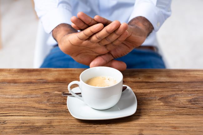 Bílý šálek s kávou na dřevěném stole, nad ním jsou překřížené mužské ruce jako náznak odmítnutí šálku s kávou.